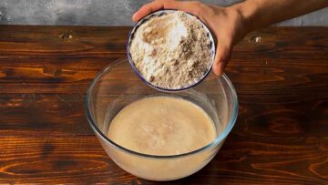 Ένα χέρι ρίχνει αλεύρι κριθαριού σε μπολ με μαγιά και νερό.